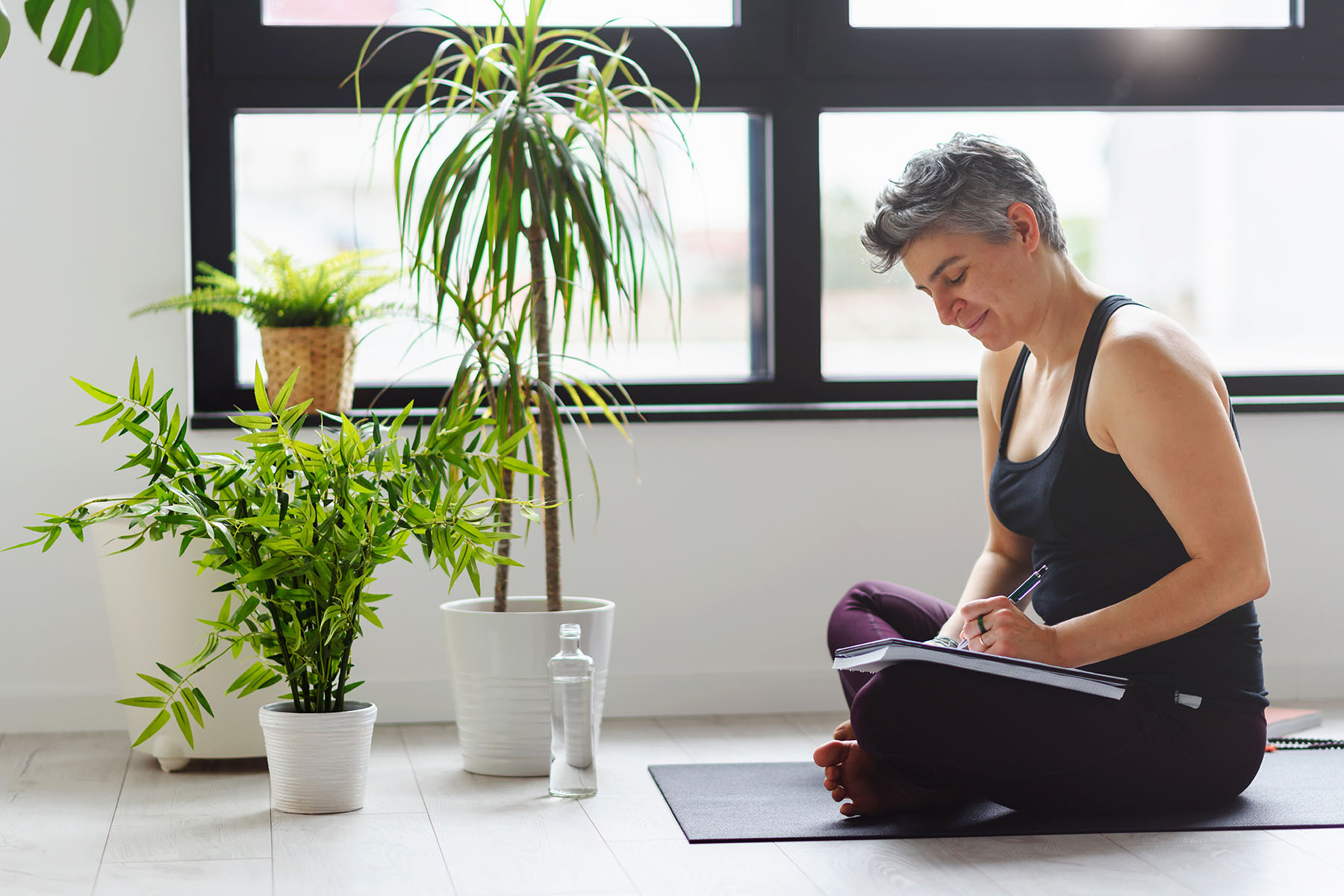 Healthy woman jots nots in a notebook while drinking water and sitting on a yoga mat.
