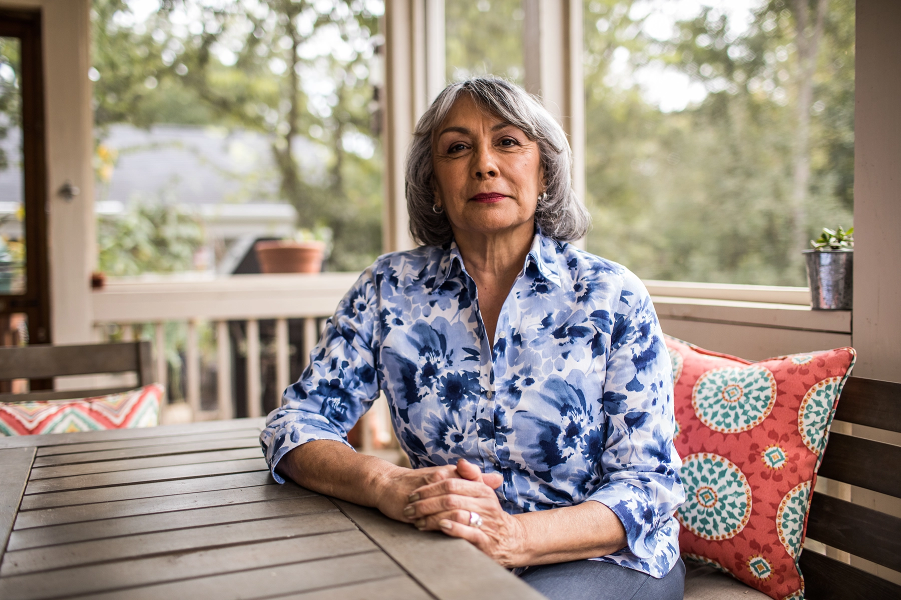 Senior Hispanic woman sits on her porch with her hands clasped.