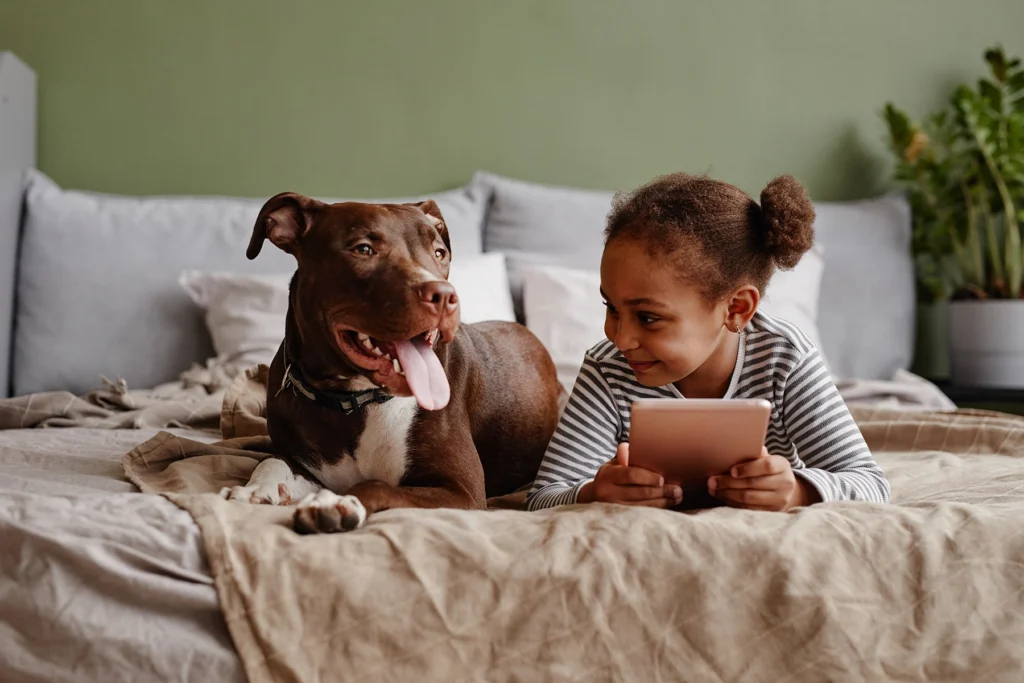 Little Black girl reads while relaxing on bed with family dog.