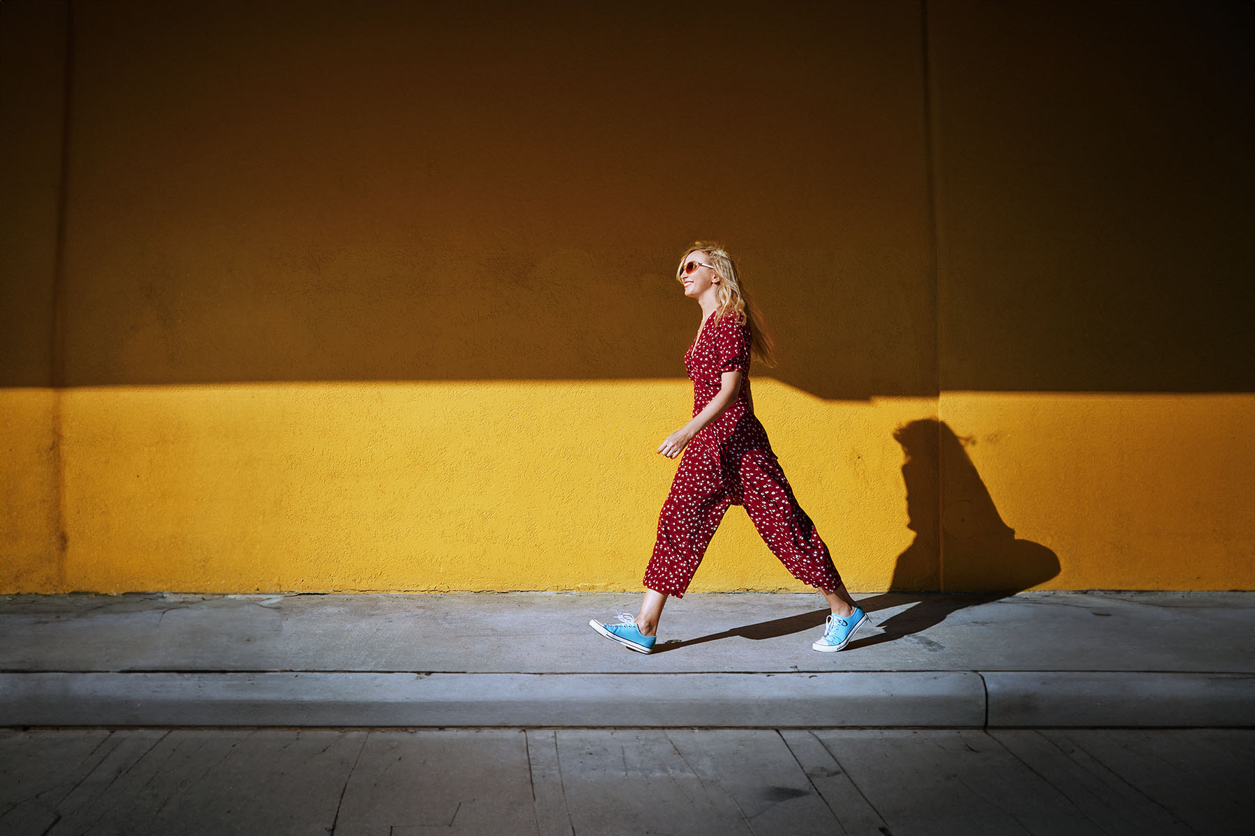 A woman in a burgundy jumpsuit and sneakers walks in front of solid yellow wall.