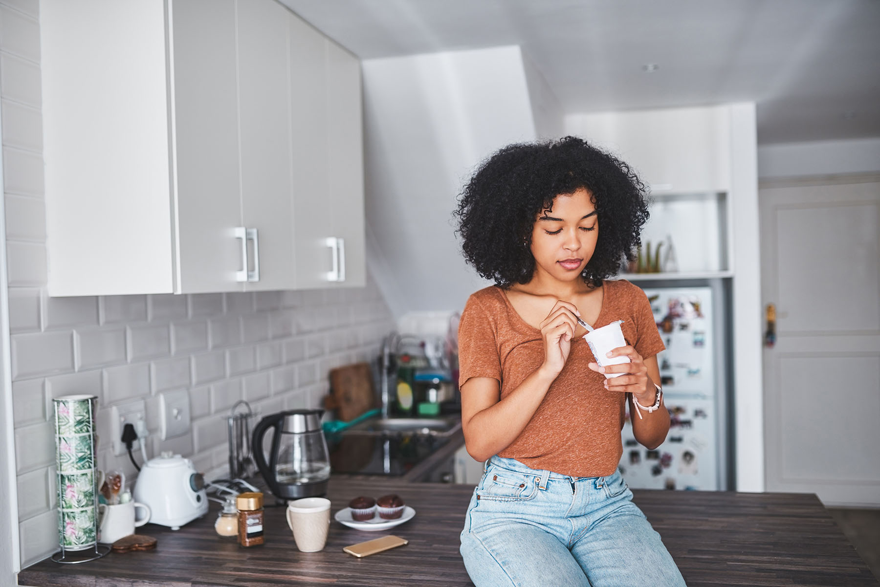Young Black woman eats yogurt while sitting on countertop.