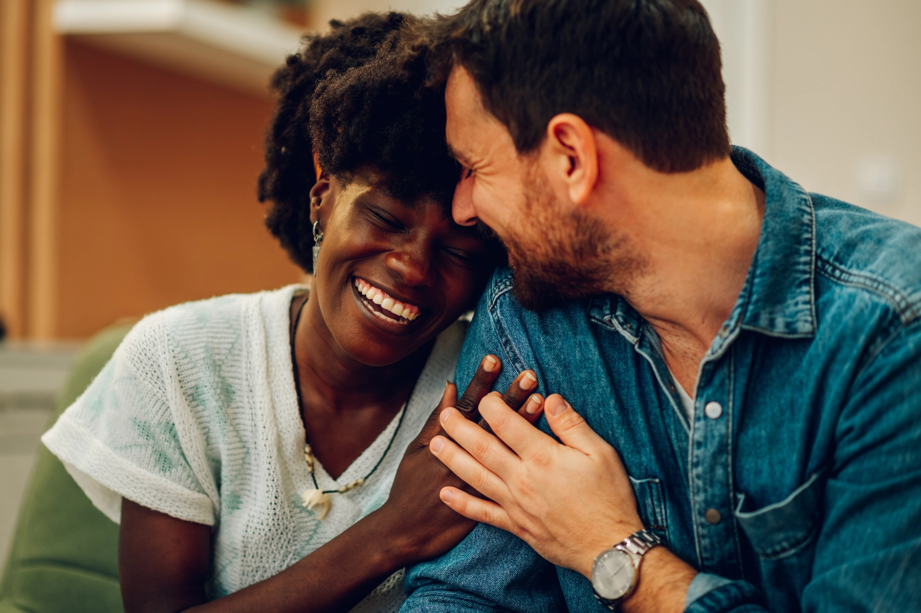 Smiling, diverse couple clasps hands and hugs on a couch.