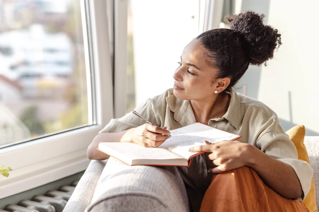 Happy, young Black woman pauses from writing in her journal to smile and look out window.