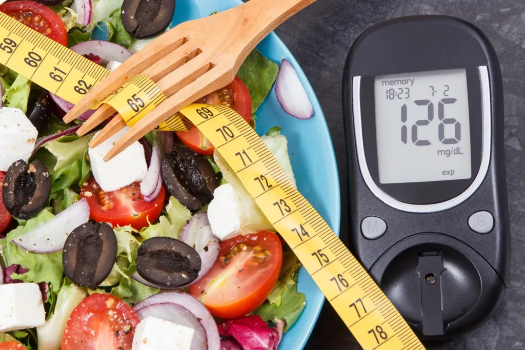 Fresh salad with lettuce, tomatoes, olives, and feta cheese, next to a measuring tape wrapped around a wooden fork and a blood glucose meter displaying 126 mg/dL, symbolizing healthy eating and diabetes management.