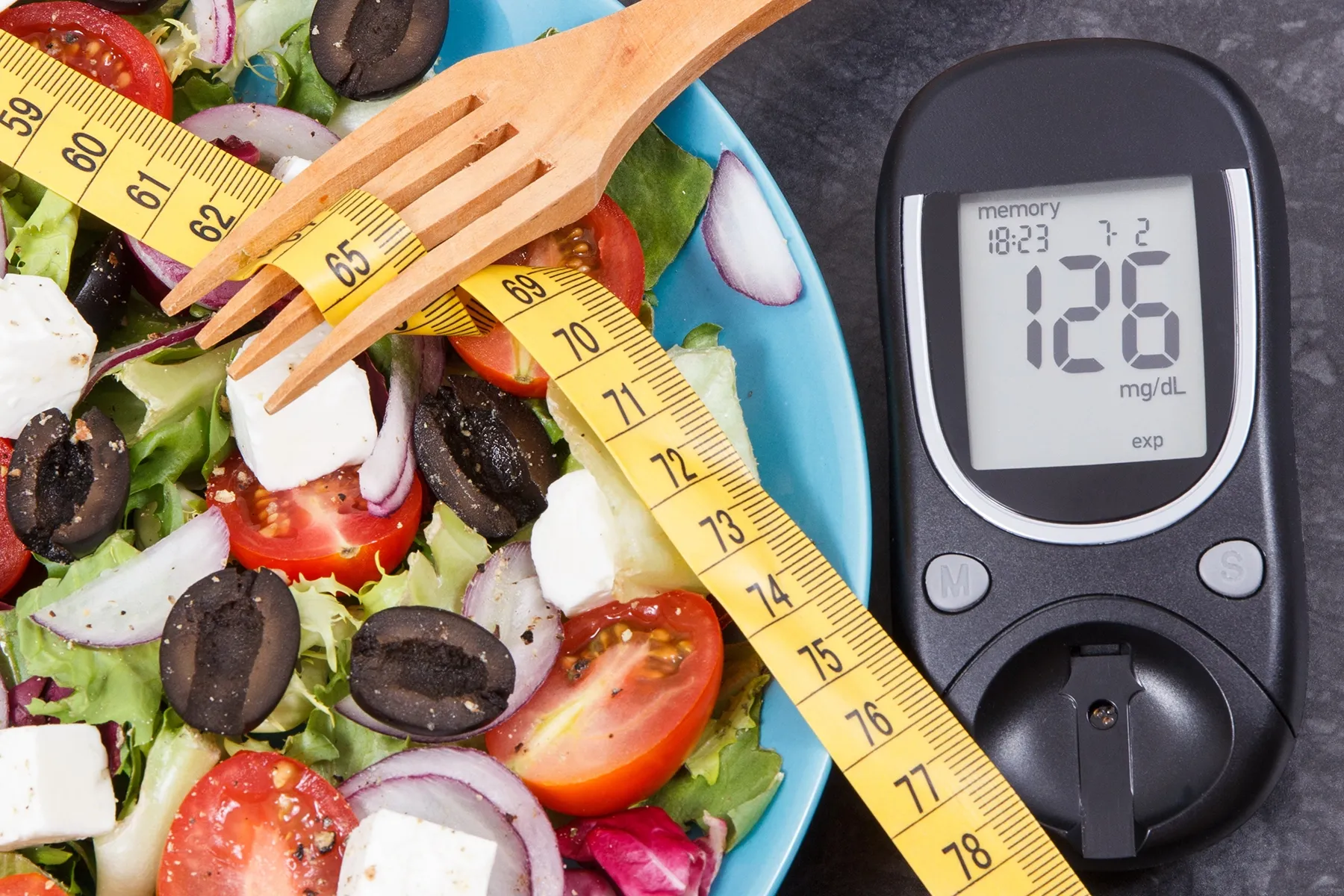 Fresh salad with lettuce, tomatoes, olives, and feta cheese, next to a measuring tape wrapped around a wooden fork and a blood glucose meter displaying 126 mg/dL, symbolizing healthy eating and diabetes management.