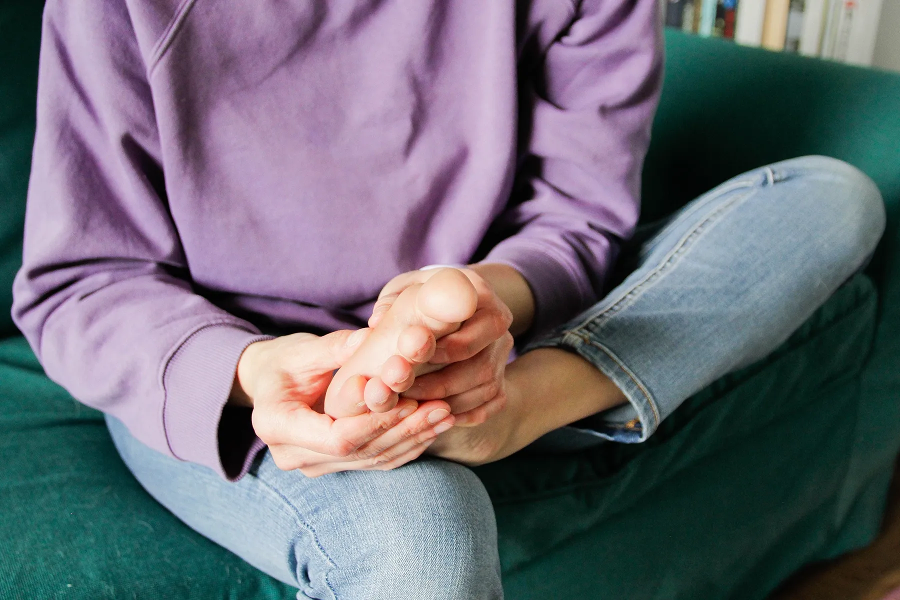 Person in a purple sweatshirt and blue jeans sitting on a green sofa, holding their bare foot.