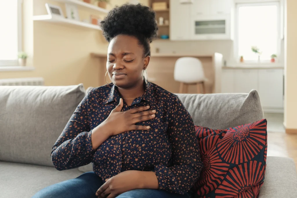 Young Black adult woman sits on her couch and holds her heart.
