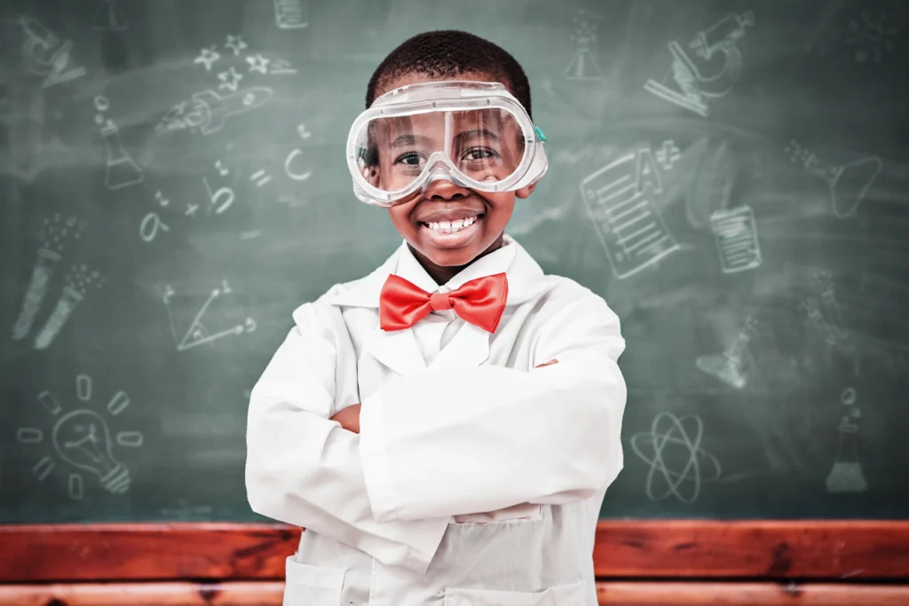 Young Black boy dressed as scientist, smiling broadly in his lab coat standing at the front of class.