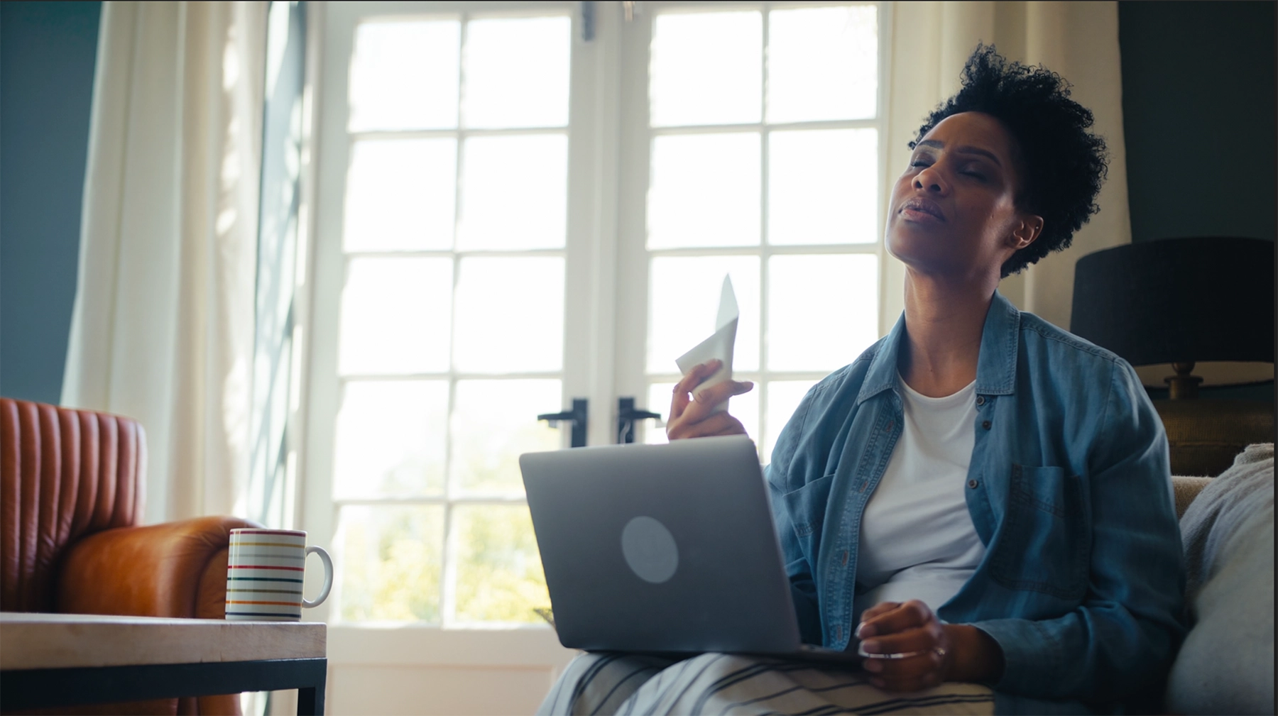 Black middle-aged woman has computer on her lap while fanning herself during perimenopause.