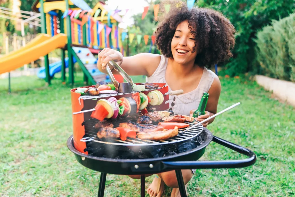 Smiling young woman with curly hair grilling meat and vegetable skewers on a charcoal barbecue. A backyard playground and colorful flags are visible in the background.