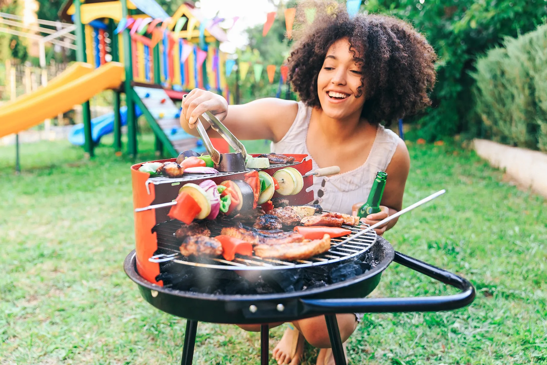 Smiling young woman with curly hair grilling meat and vegetable skewers on a charcoal barbecue. A backyard playground and colorful flags are visible in the background.