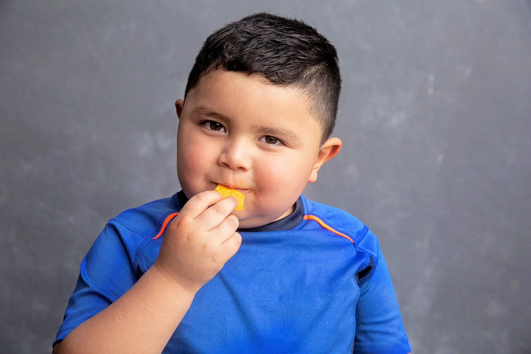Smiling young boy in a blue shirt eating a yellow chip, looking directly at the camera.