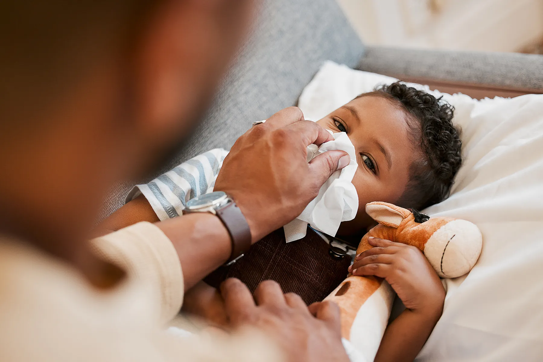 An adult's hand gently wipes the nose of a young, sick child with a tissue. The child, lying down on a white sheet, clutches a brown giraffe stuffed animal for comfort.