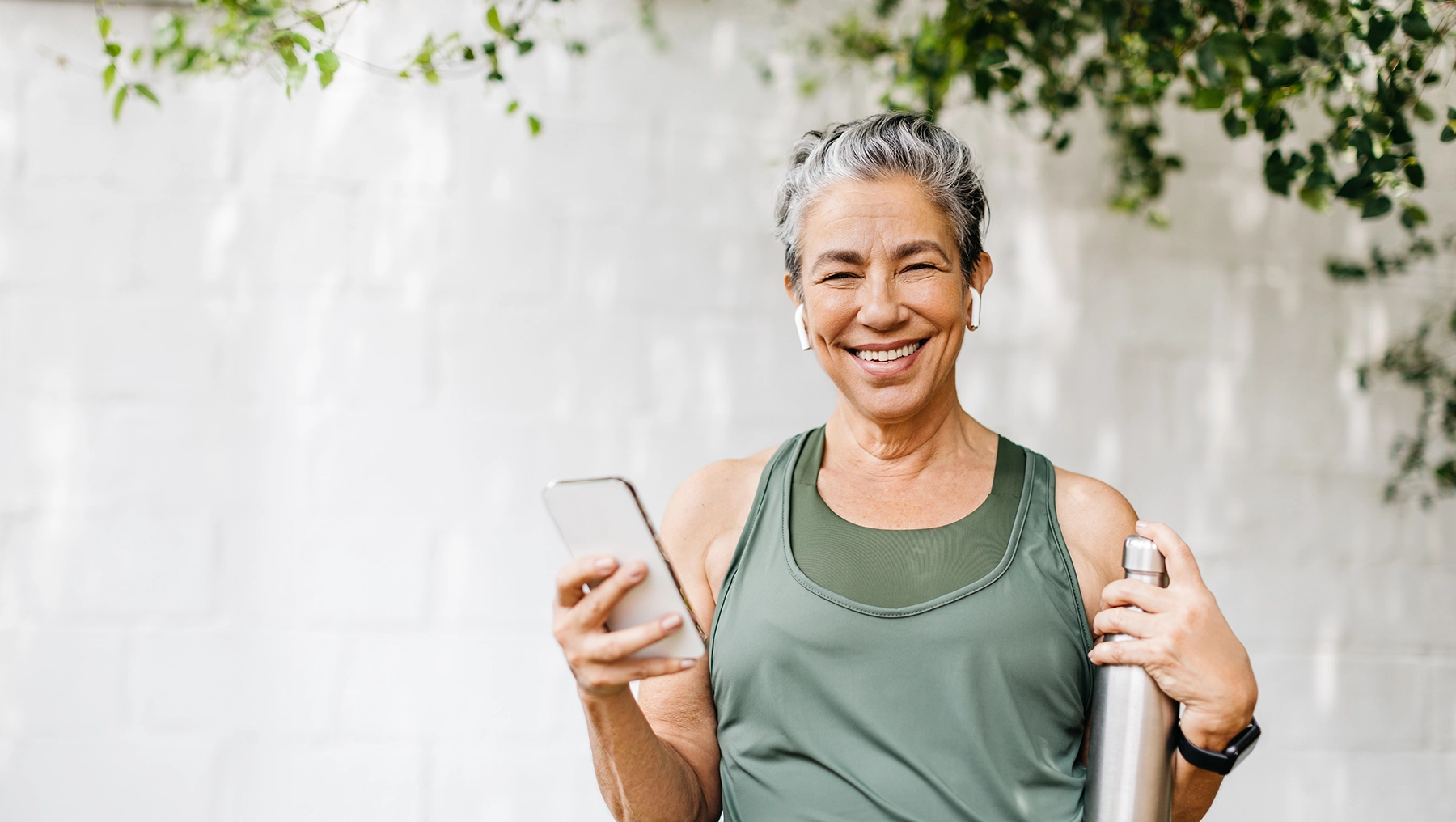 Happy, motivated, Hispanic senior woman smiles while holding a cellphone and a water bottle