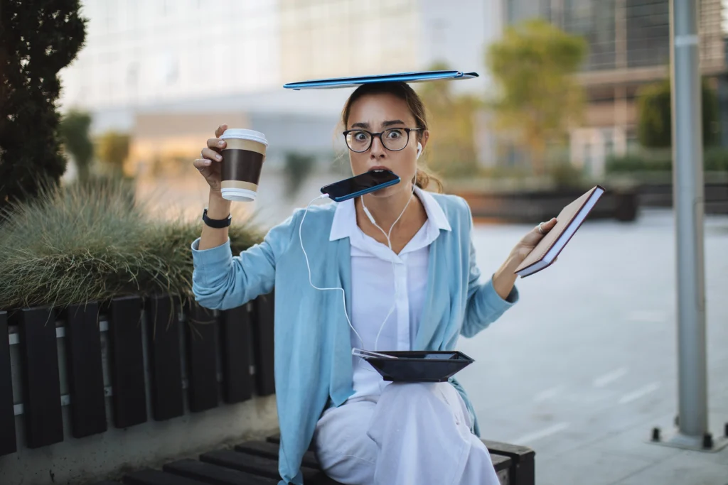 Young woman literally balances computer, phone, book and food while sitting outside. Too many distractions
