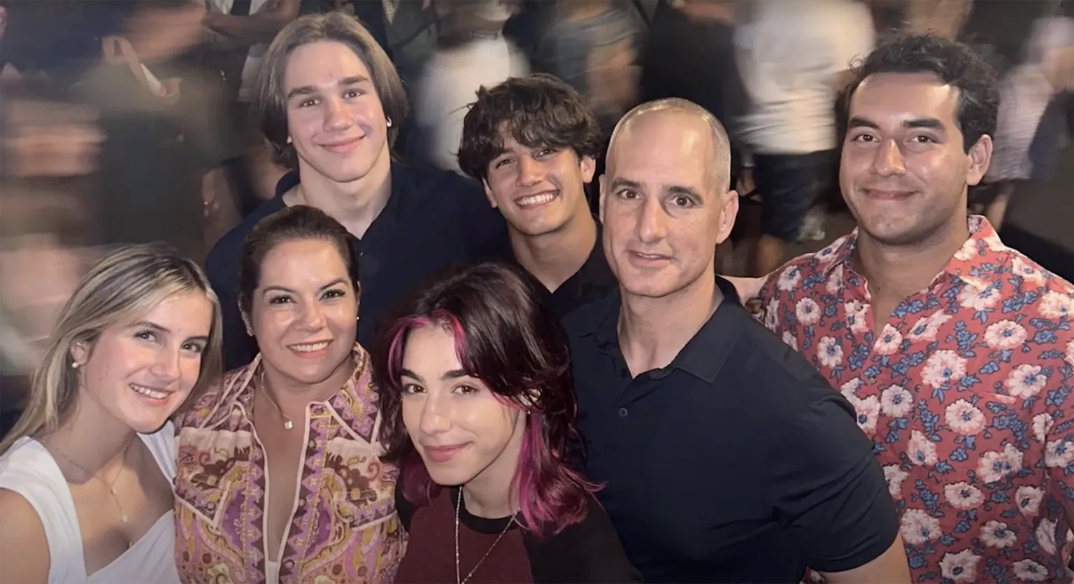 A multi-generational group of seven smiling people, three women and four men, posing closely together in an indoor setting with a blurred background of a crowd. One woman has blonde hair, another wears a patterned blouse, and a third has dark hair with pink highlights. One man wears a red floral shirt, and others are in dark shirts.