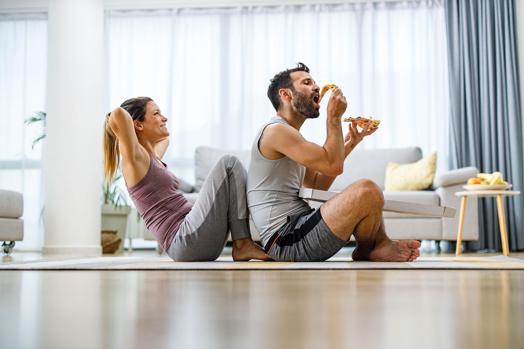 Happy woman exercising sit-ups at home while her boyfriend is assisting her and eating pizza.