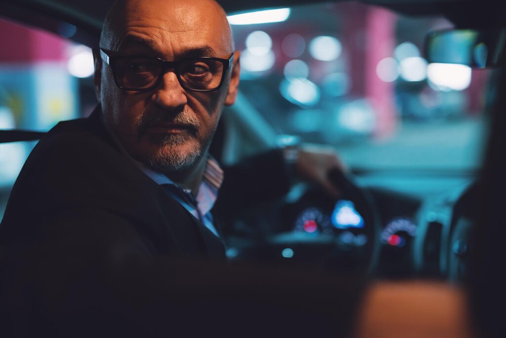 Mature man in glasses drives in reverse in a dark parking lot.