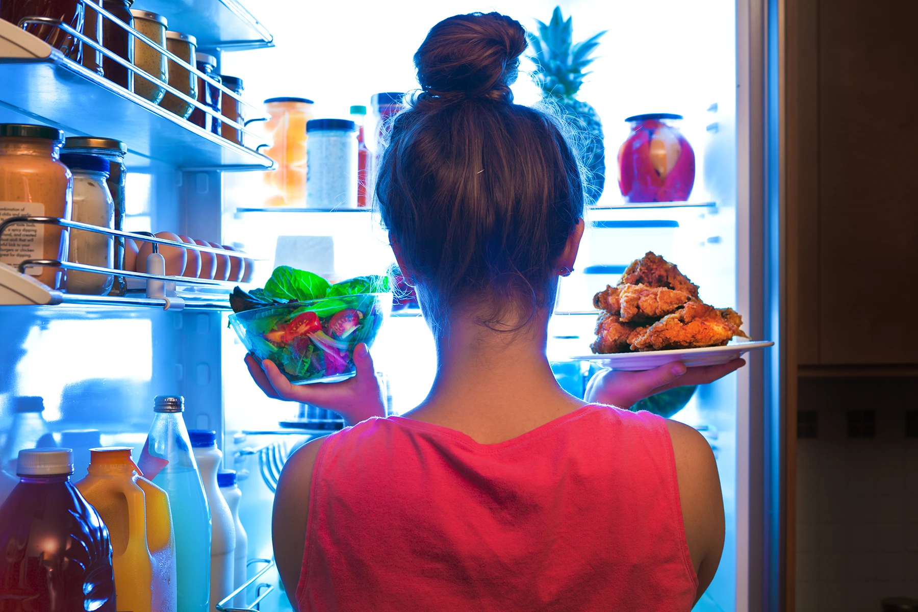 Photo of woman looking in refrigerator deciding to eat the salad or the fried chicken.