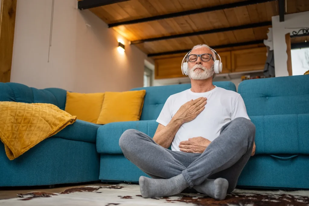 Senior man with white beard and headphones meditating cross-legged on a rug in a cozy living room with a teal sofa and yellow pillows.