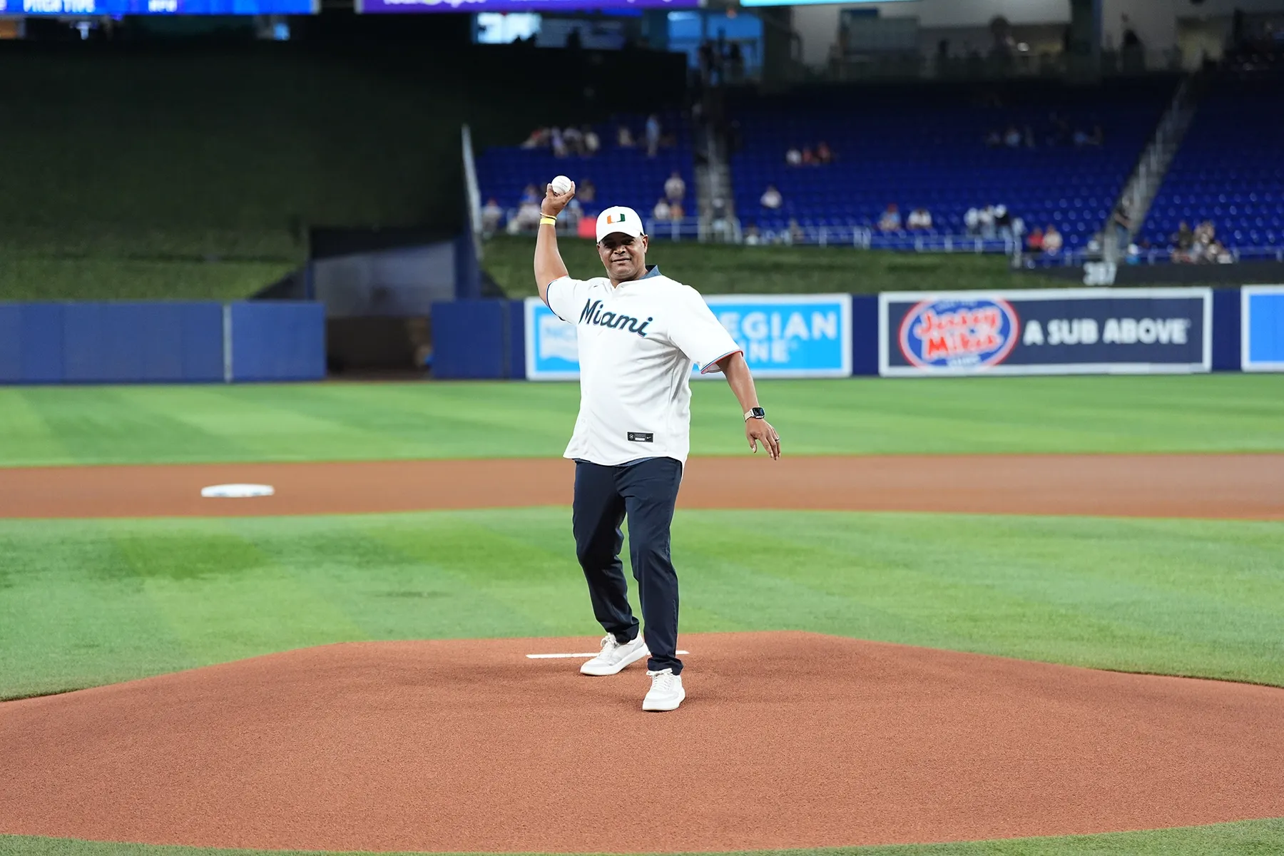 An older man in a white "Miami" baseball jersey and a "U" logo hat delivers a pitch from the pitcher's mound at a stadium, with blue seats and sponsor banners in the background.