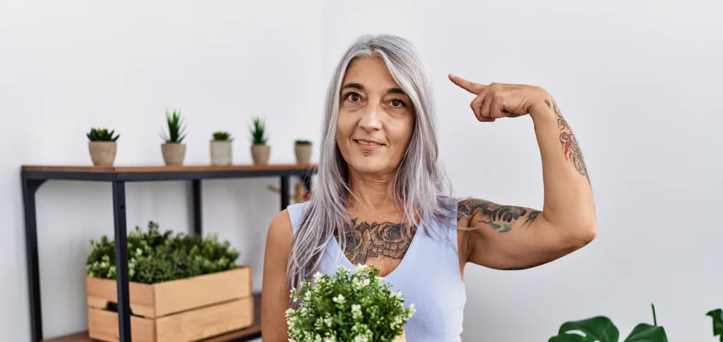 Middle aged Hispanic woman holds plant and points at her gray hair.