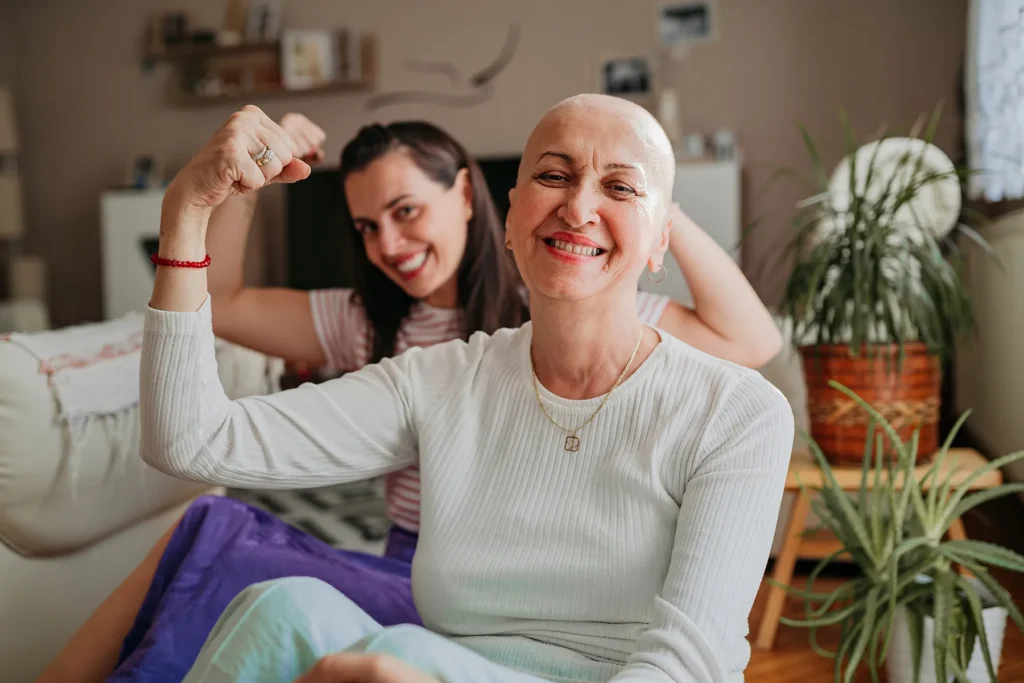 A bald woman and a younger woman smiling broadly and flexing their biceps, symbolizing strength, resilience, and mutual support in a home environment.