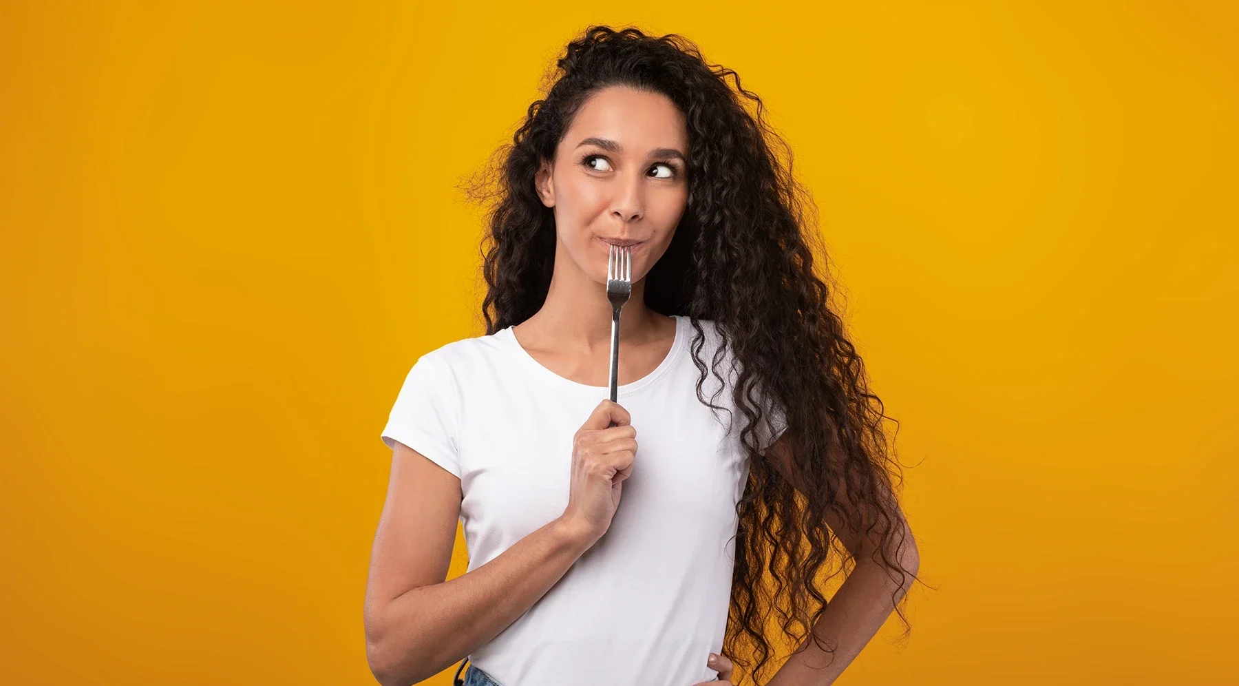 Woman in white t-shirt looks sideways while biting on a fork, eagerly awaiting food.