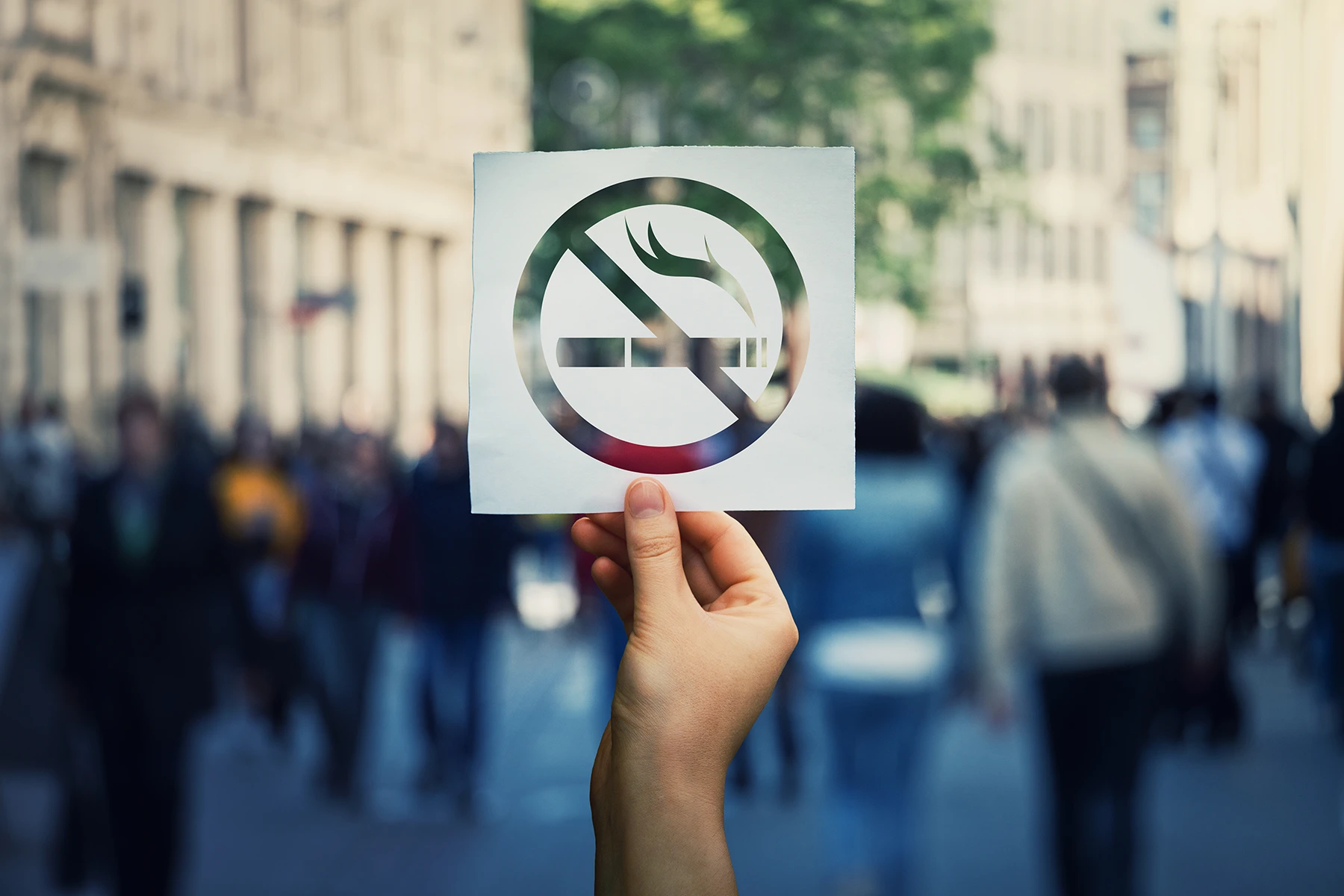 Hand holds up a paper no smoking sign on a busy city street.