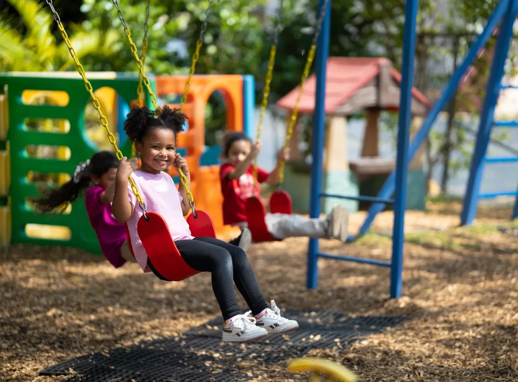 A smiling young girl with dark curly hair swings joyfully on a red seat at an outdoor playground, wearing a pink shirt and black leggings. Two other children are also on swings in the blurred background, with colorful play structures and wood chips on the ground.