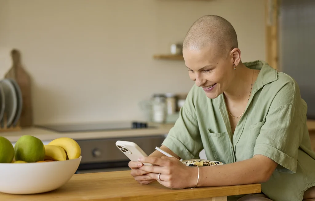 Female cancer patient smiles at her phone while leaning on kitchen counter.