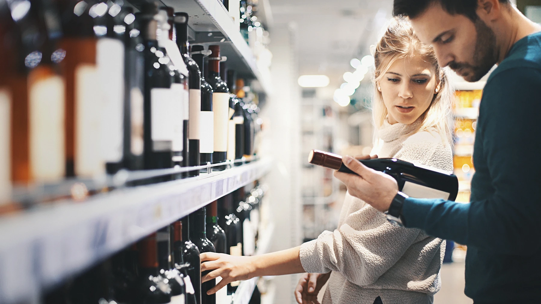 A couple reads the label on a bottle of wine while shopping at a supermarket.