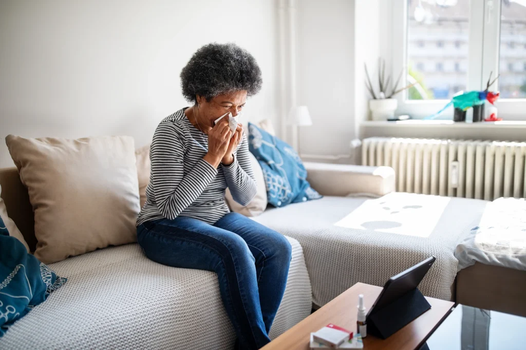 Woman sits on couch blowing her nose while on a video call.