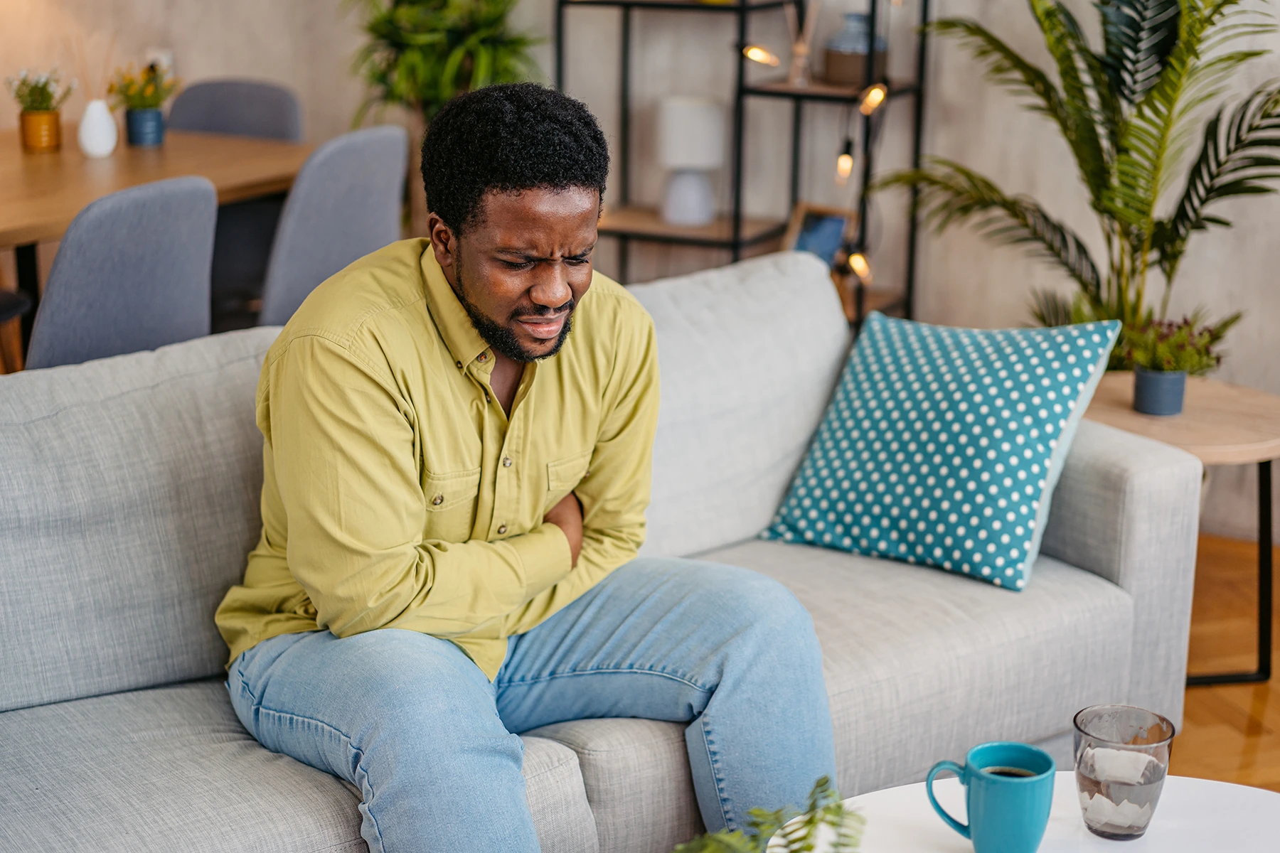 A young Black man sits on a couch gripping his stomach due to an h.pylori infection.