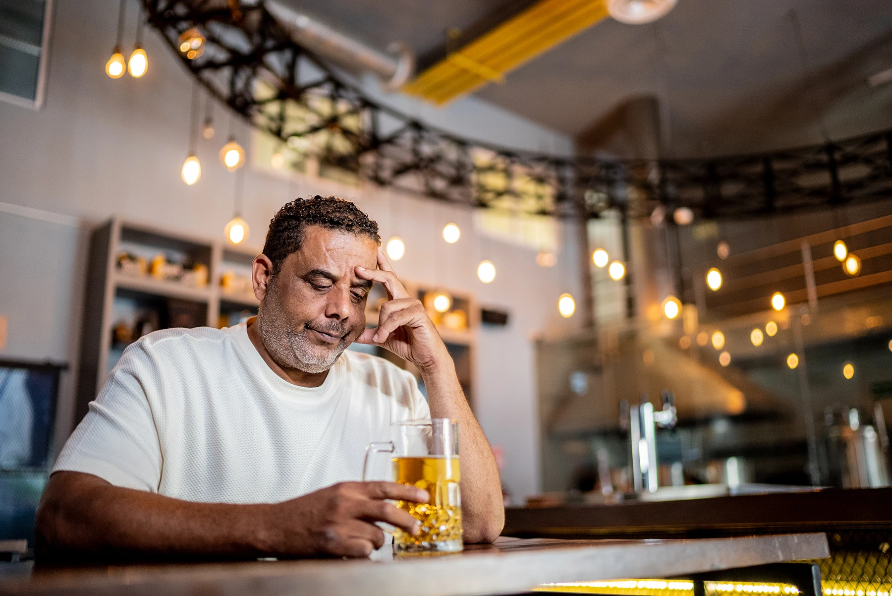 Hispanic man sits at bar, thinking while drinking a beer.