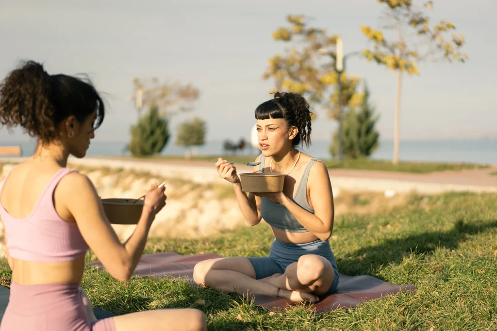 Two young women sit outside eating a protein and fiber rich salad after a yoga class in the park.