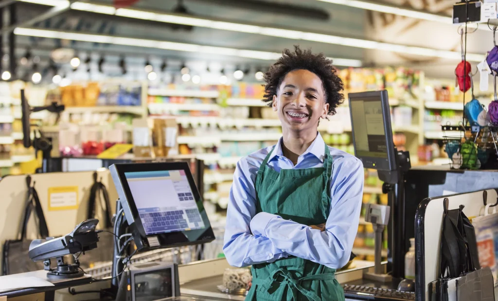 Young teen boy smiles widely working as a cashier at a grocery store.