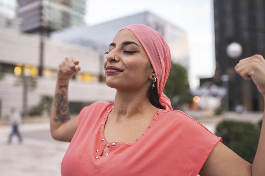 Young woman in a headscarf flexes while walking outside in a city. Stock image to show cancer patient feeling strong.