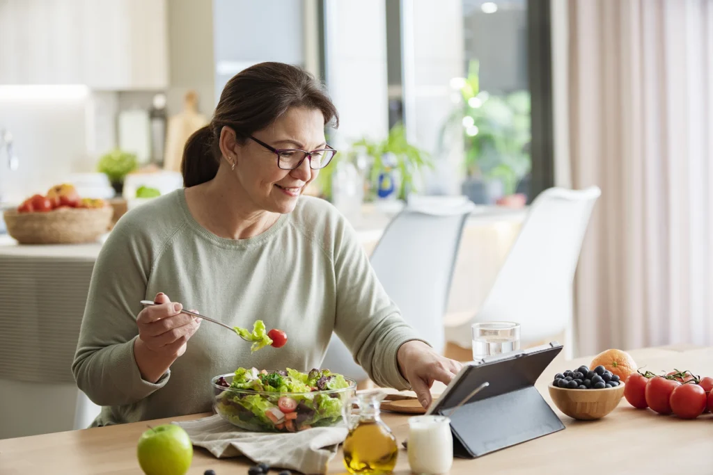 Woman is sitting at the table using digital tablet while having healthy vegetables meal.