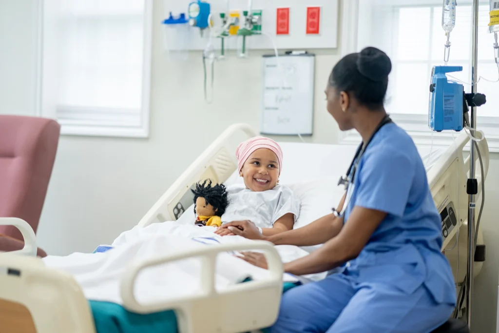 A female nurse of African decent sits with a young Cancer patient as she tries to cheer her up. The little girl is wearing a head scarf to keep her warm and is hugging a stuffed doll for comfort.