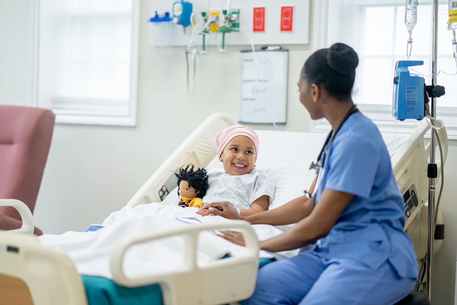 A female nurse of African decent sits with a young Cancer patient as she tries to cheer her up. The little girl is wearing a head scarf to keep her warm and is hugging a stuffed doll for comfort.
