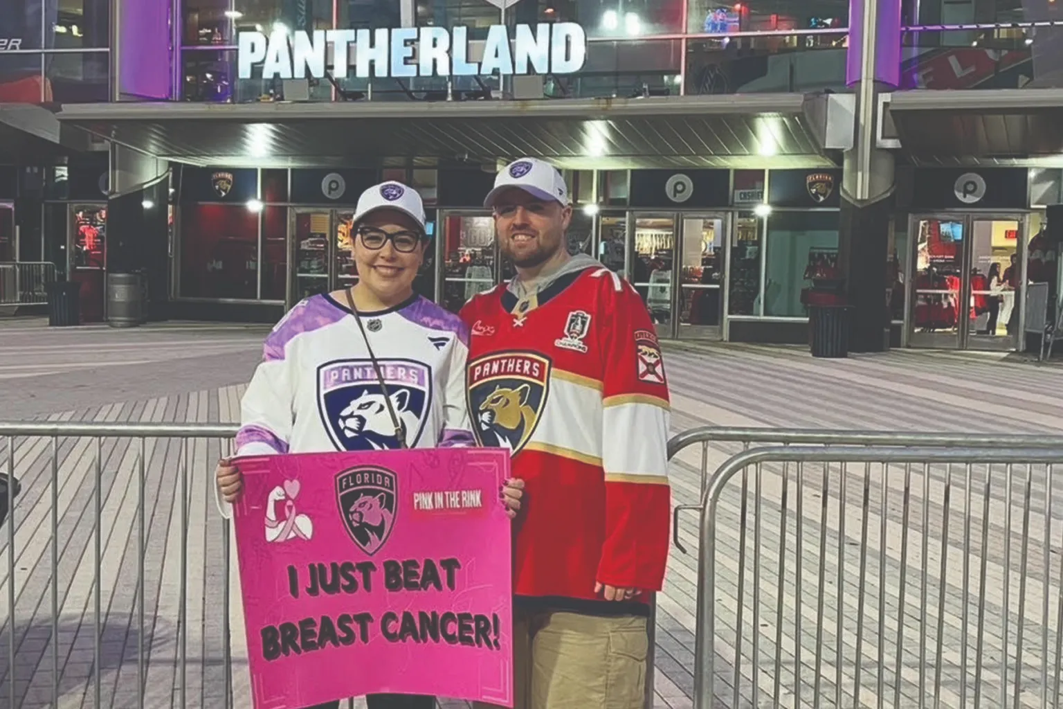 Smiling woman in a Florida Panthers jersey holding a pink "I JUST BEAT BREAST CANCER!" sign, posing with a man in a red Panthers jersey outside the "Pantherland" arena during a "Pink in the Rink" event.