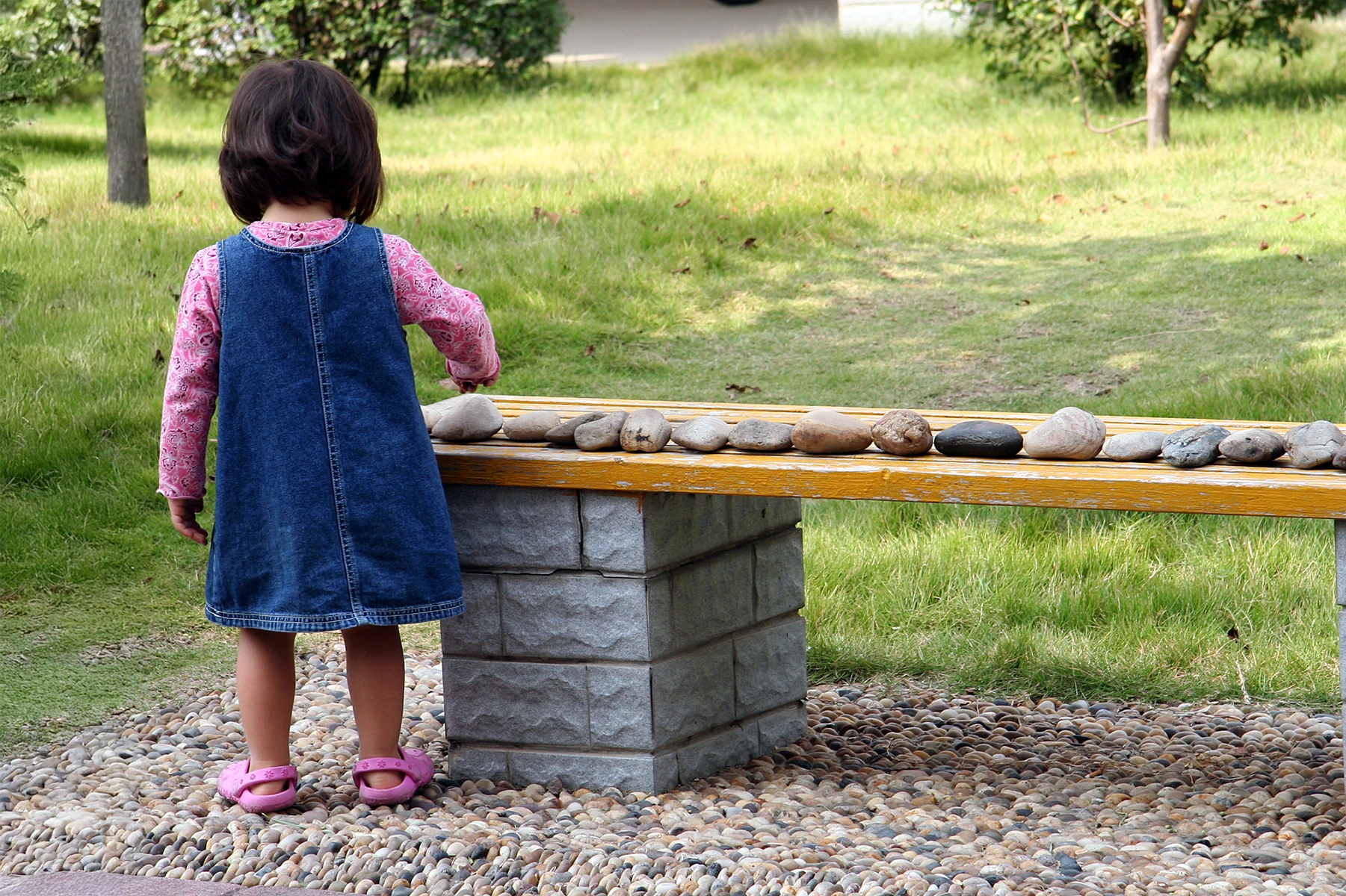 Little girl in demin dress lines up rocks in a perfect line on bench.