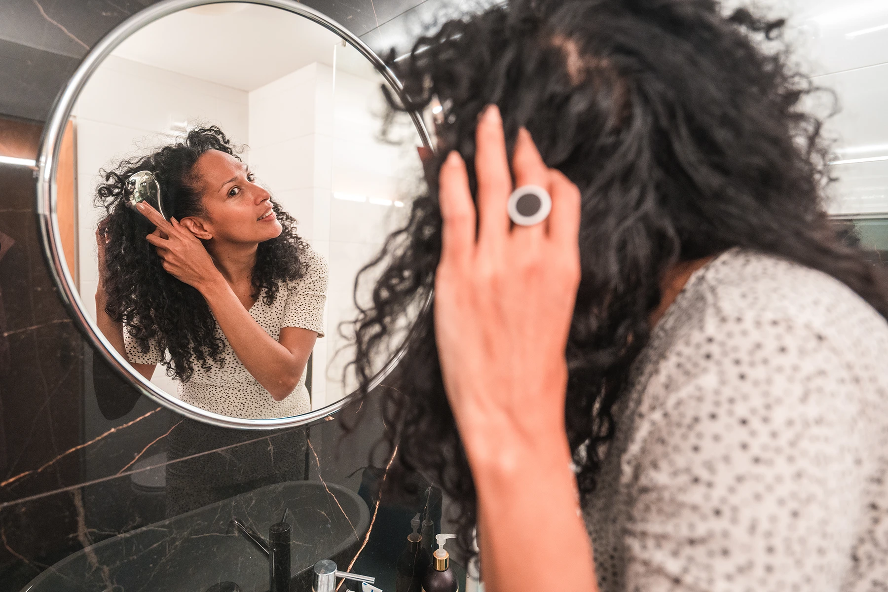 Hispanic woman brushes her hair in mirror, showing slight hair loss due to life changes. Stock image.