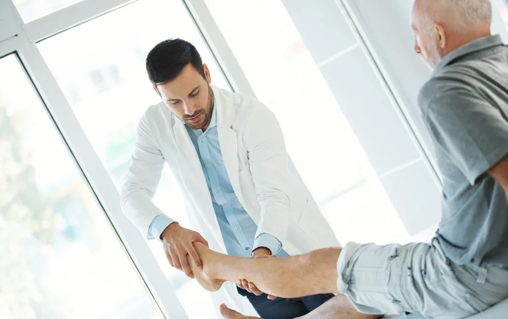 Male doctor in a white coat performing a physical leg examination on a seated older male patient in a bright, modern medical clinic.