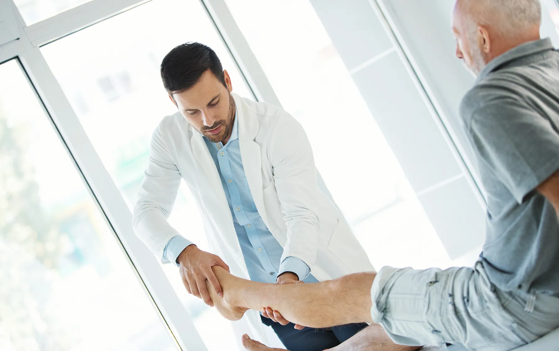 Male doctor in a white coat performing a physical leg examination on a seated older male patient in a bright, modern medical clinic.