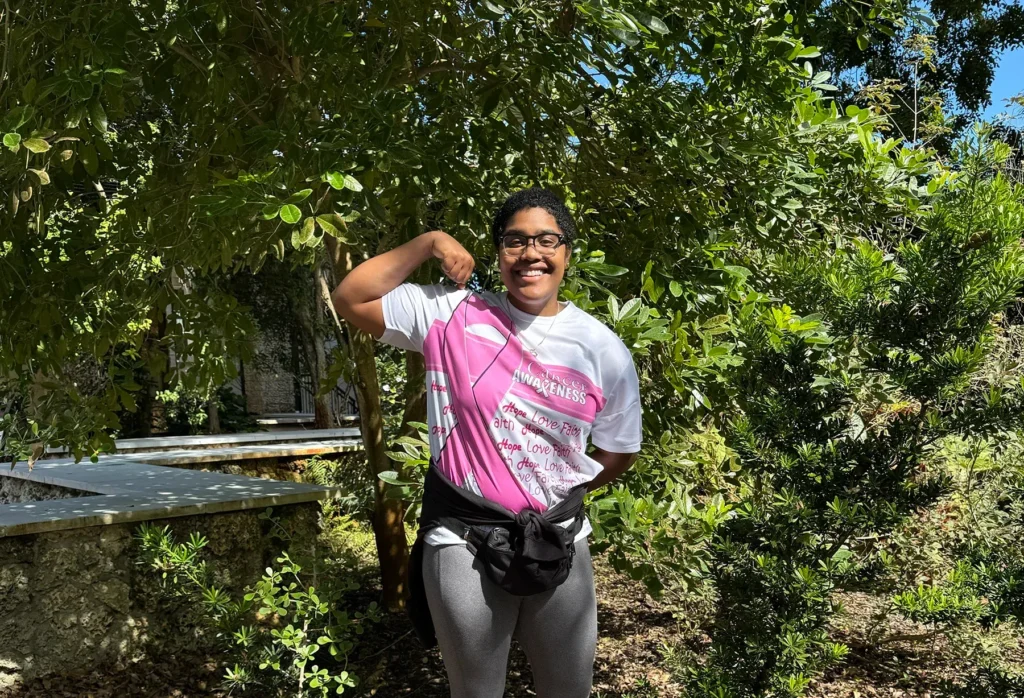 Smiling young Black person with short curly hair and glasses, wearing a white t-shirt with a large pink breast cancer awareness ribbon and "Cancer Awareness" text, flexing their arm. They stand outdoors in a lush green garden under bright sunlight.