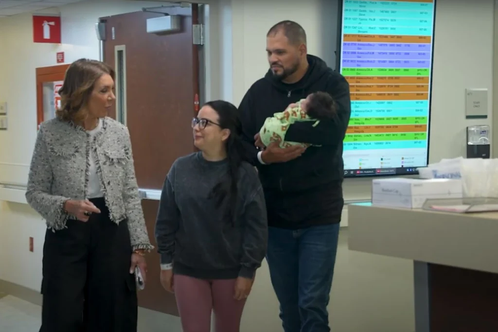 Three adults walk through a hospital hallway, one carrying a baby wrapped in a blanket. A large digital screen displays colored rows of text, possibly patient schedules. Medical equipment is visible on a counter to the right.