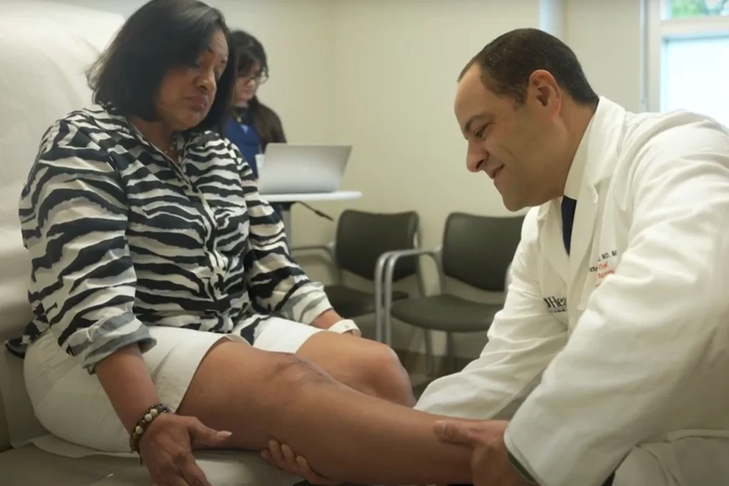 A healthcare professional in a white coat evaluates a patient's leg while seated on an exam table. The patient wears a striped shirt and white shorts. In the background, a person works on a laptop near a row of chairs.