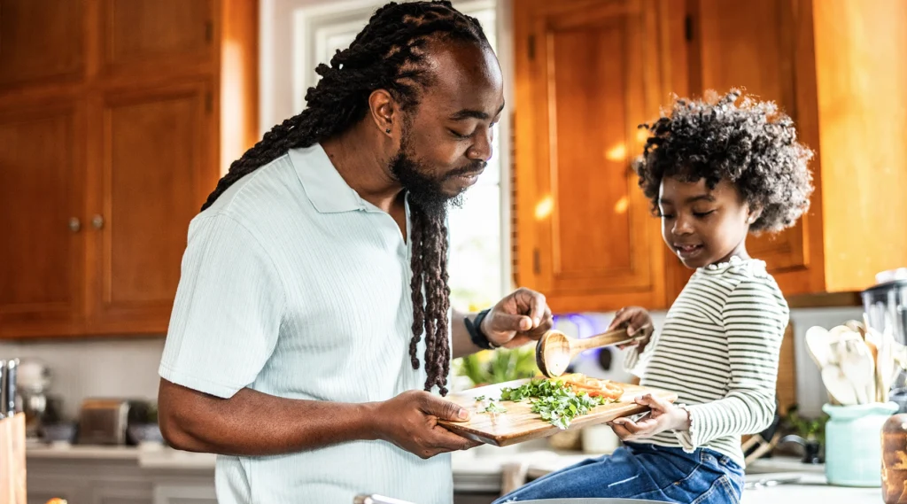 Good lifestyle choices help prevent cancer. Photo of Black father happily preparing a healthy dinner with his adorable toddler daughter in the kitchen.